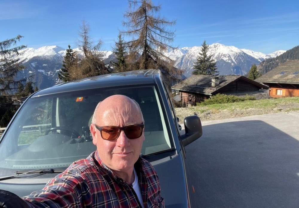 A selfie in front of the Able Transport van with snow-capped Swiss Alps and wooden chalets behind